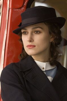 a woman wearing a black hat and coat leaning against a red train car with her arms crossed