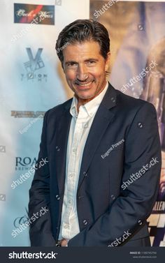 a smiling man in a suit and white shirt stands on the red carpet at an event