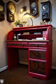 an old fashioned red desk with clocks on the wall behind it and a lamp in front of it