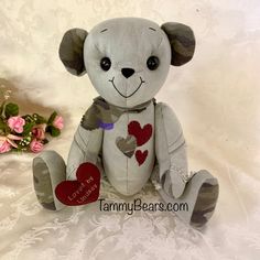 a gray teddy bear with hearts on it's chest sitting next to pink flowers