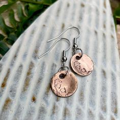 two metal discs with flowers on them are hanging from silver earwires, sitting on top of a stone slab