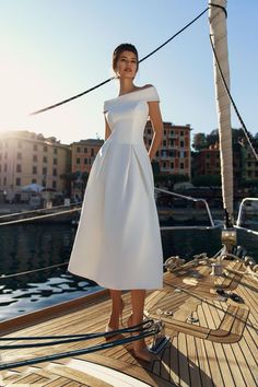 a woman in a white dress standing on the deck of a sailboat with buildings in the background