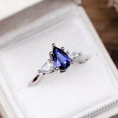 a ring with three pear shaped diamonds and a blue tear droplet sits in a white box