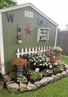 a small garden shed with flowers in the front and on the side, sitting next to a white picket fence