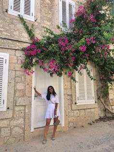 a woman standing in front of a door with pink flowers growing on the side of it