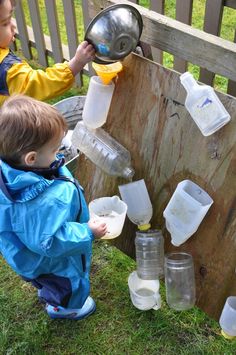 two children are playing with plastic jugs on the grass near a wooden bench and fence