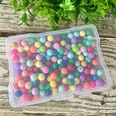 a plastic container filled with lots of colorful beads next to some green leaves on a wooden table