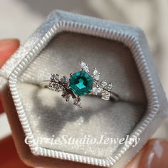 a person holding an engagement ring in a box with the top section open to show it's green stone and diamond accents