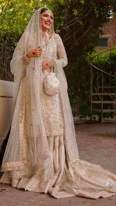 a woman in a white dress and veil standing next to a car with trees behind her