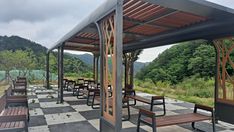 an outdoor seating area with wooden benches and tiled flooring, surrounded by green trees