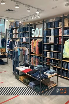 Interior da loja da marca de roupas Levi’s, no Shopping Riomar, em Recife. Araras dispostas junto as paredes e distribuídas pelo salão da loja apresentam os produtos da marca, a maioria em tecido jeans, alguns pendurados em cabides e outros dobrados em estantes. Os móveis de apoio, caixa, portas do provador são todos em madeira e com toques industriais. Todo o ambiente é muito bem iluminado. Men's Clothing Store Design, Clothing Shop Interiors, Clothing Store Interior Design, Fashion Shop Interior, Retail Store Layout, Store Shelves Design