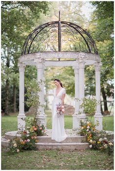 a woman standing in front of a gazebo with flowers around her and holding a bouquet