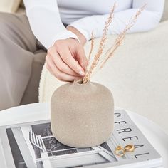 a person sitting on a couch with their hands in a small round stone vase that is placed on top of a magazine