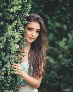 a beautiful young woman standing next to a lush green bush covered in leaves and looking at the camera