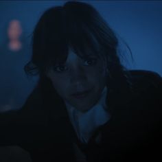 a woman with dark hair and blue eyes looks at the camera in a darkened room