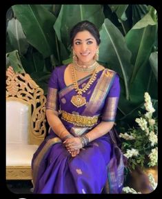 a woman in a purple and gold sari sitting on a bench with greenery behind her