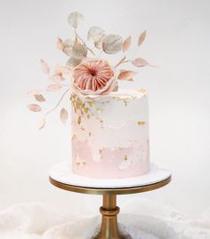 a white and pink cake with gold leaf decoration on a small metal stand in front of a white wall