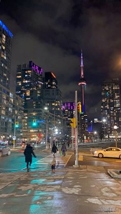 people are walking on the sidewalk in front of some tall buildings at night with bright lights