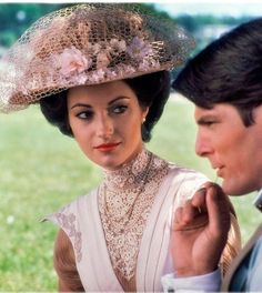 a man standing next to a woman in a dress and hat on top of a grass covered field