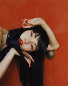 a woman with long black hair and red lipstick posing for a photo in front of an orange wall