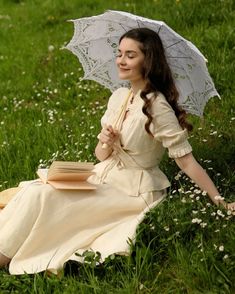 a woman sitting in the grass with an umbrella over her head, reading a book