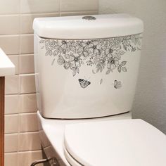 a white toilet sitting next to a sink in a bathroom under a window with flowers and butterflies on the lid