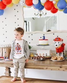 a little boy standing in front of a table with cake and balloons on it's wall