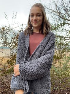 a woman standing in front of some bushes wearing a gray knitted sweater and jeans