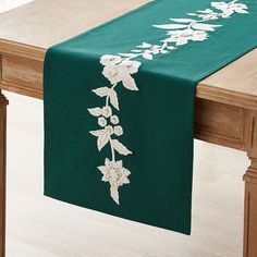 a green table runner with white flowers and leaves on the edge is sitting on a wooden table