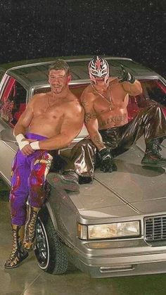 two men sitting on the hood of a car in front of another man wearing wrestling gear