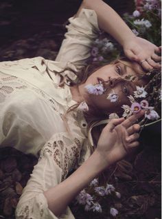 a woman laying on the ground with flowers in her hand and holding something up to her face