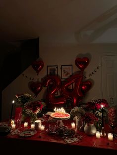 a red table topped with a cake covered in candles and heart shaped balloons next to a number twenty sign