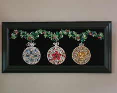 three ornaments are hanging on the wall in front of a black framed art piece with holly and red berries