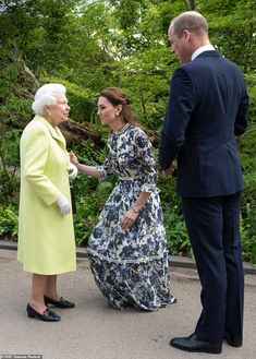 two men and a woman standing next to each other in front of green trees, talking