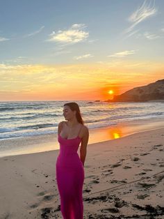 a woman in a long pink dress standing on the beach at sunset with her arms behind her back