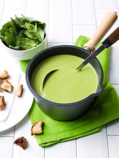 a green soup in a pot with bread and spinach on the side next to it