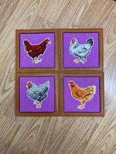 four cross stitch chickens on purple squares with brown trim around the edges, sitting on a wooden surface