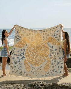 two young women holding up a yellow and white blanket on the beach near the ocean