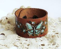 "A soft camel brown leather cuff decorated with a large butterfly stud, accented in turquoise blue and surrounded by tiny painted blue flowers sprinkled among golden-green leaves! A few select flowers sparkle with the help of little blue Swarovski crystal rhinestones for a touch of glam! The cuff is sure to fit most wrists as it is adjustable with two sturdy snap size options! An artful addition to your wardrobe! Adjustable Soft Leather Cuff, 9\" long, 1 1/2\" wide with two snap size options, wi Adjustable Brown Jewelry For Spring, Brown Jewelry For Spring Gift, Spring Brown Jewelry Gift, Spring Gift Brown Jewelry, Fall Pillar Candles, Hand Painted Butterfly, Painted Butterfly, Hand Painted Candles, Golden Green