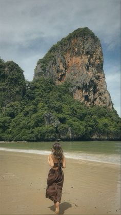 a woman walking on top of a sandy beach next to the ocean with mountains in the background