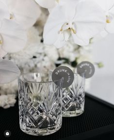 two crystal tumblers sitting next to each other on a table with flowers in the background