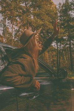 a woman with long hair wearing a hat leaning out the window of a car and waving