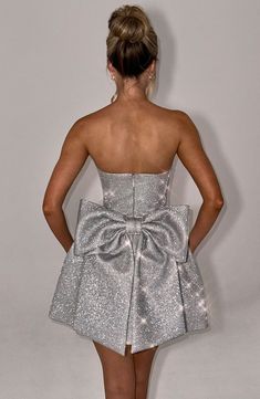 THE dress of the season has landed and you're going to be obsessed. Meet Alice. our dreamy sparkly mini with a pretty bubble shaped skirt and waist cinching bodice. This strapless style is complete with an oversized bow to the back for perfect feminine drama.   Colour: Silver Sparkle. Premium non-stretch glitter woven fabric. Fully lined. Strapless neckline. Waist cinching. Detachable. oversized bow detail to back. Voluminous bubble shaped skirt. Lace up to the reverse. Invisible zipper. Mini length. Model is an XS and is wearing an XS. Dresses With Bows In The Back, Sparkle Short Dress, Vegas Ootd, Homecoming Dresses Corset, Singer Dr, Midi Dress Wedding Guest, 17 Birthday, Bow Mini Dress, Formal Wear Women