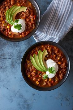 two bowls filled with chili and avocado on top of a blue countertop