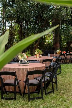 an outdoor table set up with black chairs and round wooden tables surrounded by greenery