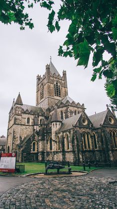 an old church with a bench in front of it and trees around the building, on a cloudy day