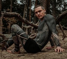 a man sitting on the ground in front of some trees wearing black and white tattoos