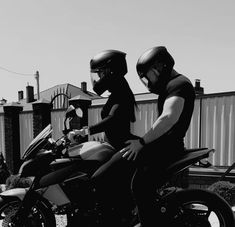 black and white photograph of two people wearing helmets sitting on a motorbike in front of a fence