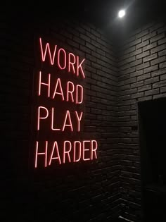 a red neon sign that says work hard play harder on a brick wall in a dark room