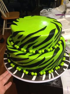 a green cake with black and white designs on it sitting on a table next to a person's hand